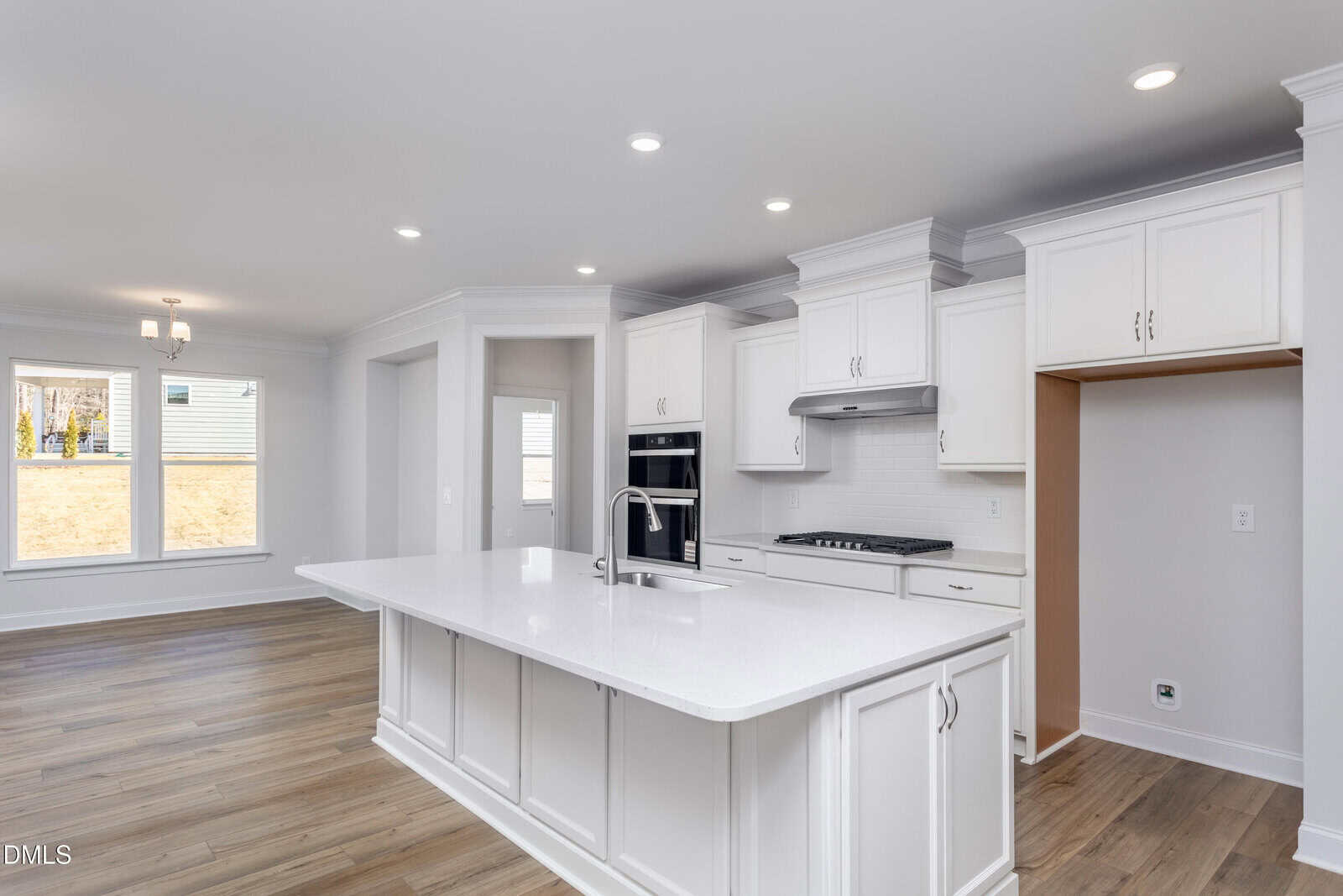 Bright, modern kitchen with white cabinets, a large island with sink, built-in oven, gas cooktop, and open area leading to a dining space with large windows and wood flooring throughout.
