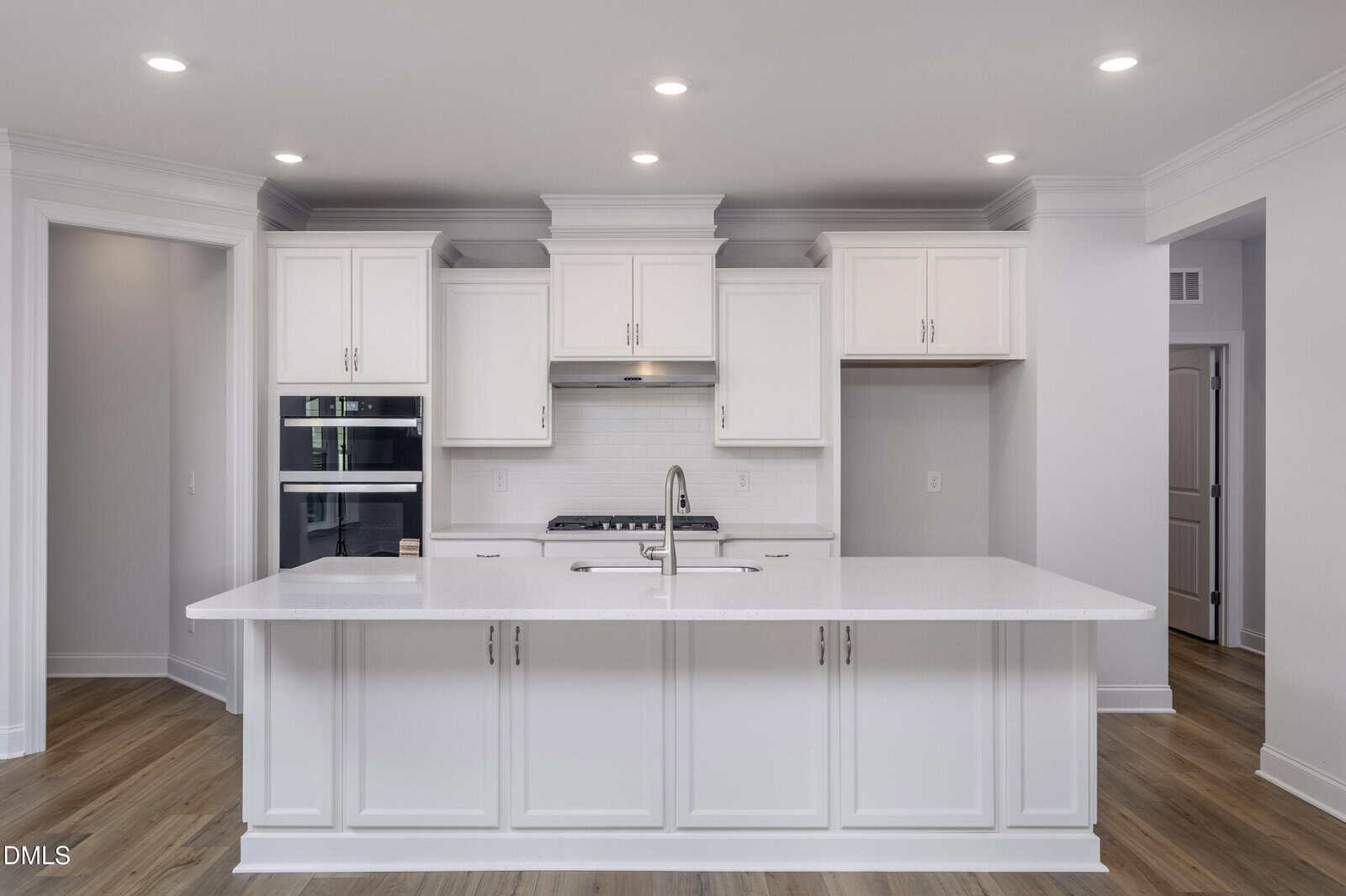 Modern kitchen with white cabinets, stainless steel appliances, a large island with a built-in sink, and light wood flooring, all illuminated by recessed ceiling lights.