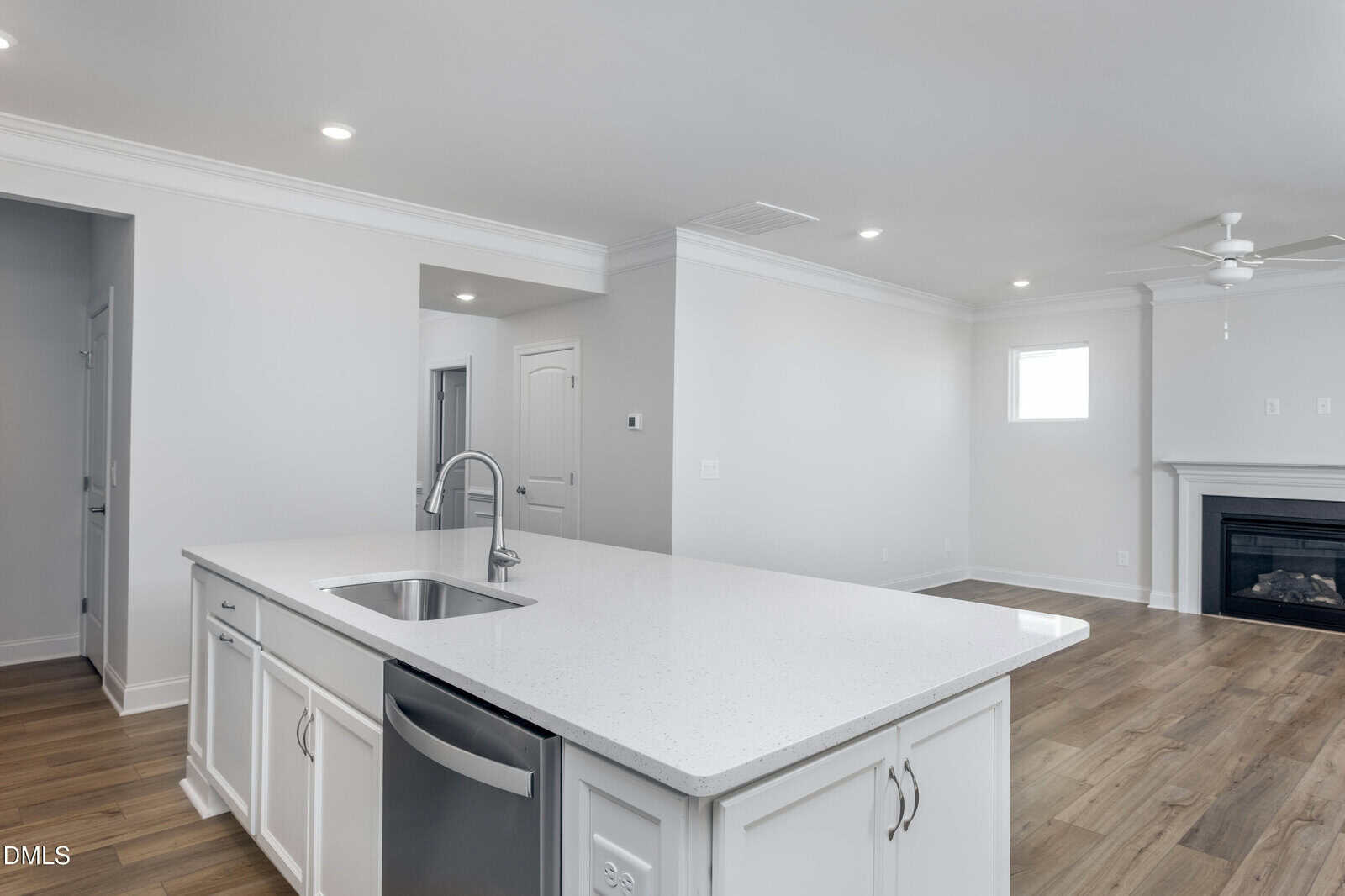 Modern, open-concept kitchen and living area with white cabinets, a large island with a sink, stainless steel appliances, light wood flooring, a ceiling fan, and a fireplace against a white wall.