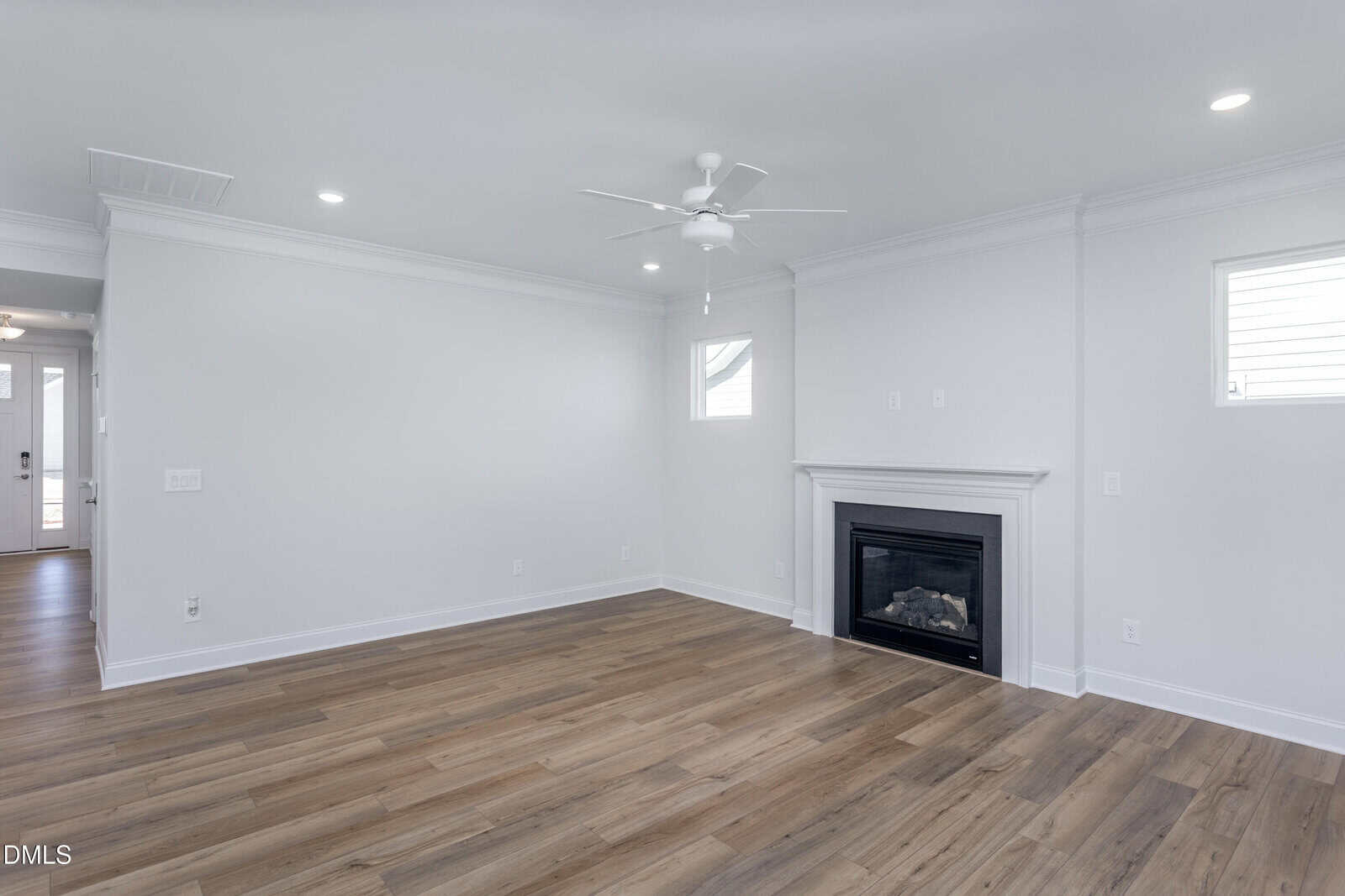 A bright, empty living room with light-colored walls, wood flooring, recessed lighting, a ceiling fan, two small windows, and a white fireplace. An entryway with a front door is visible in the background.
