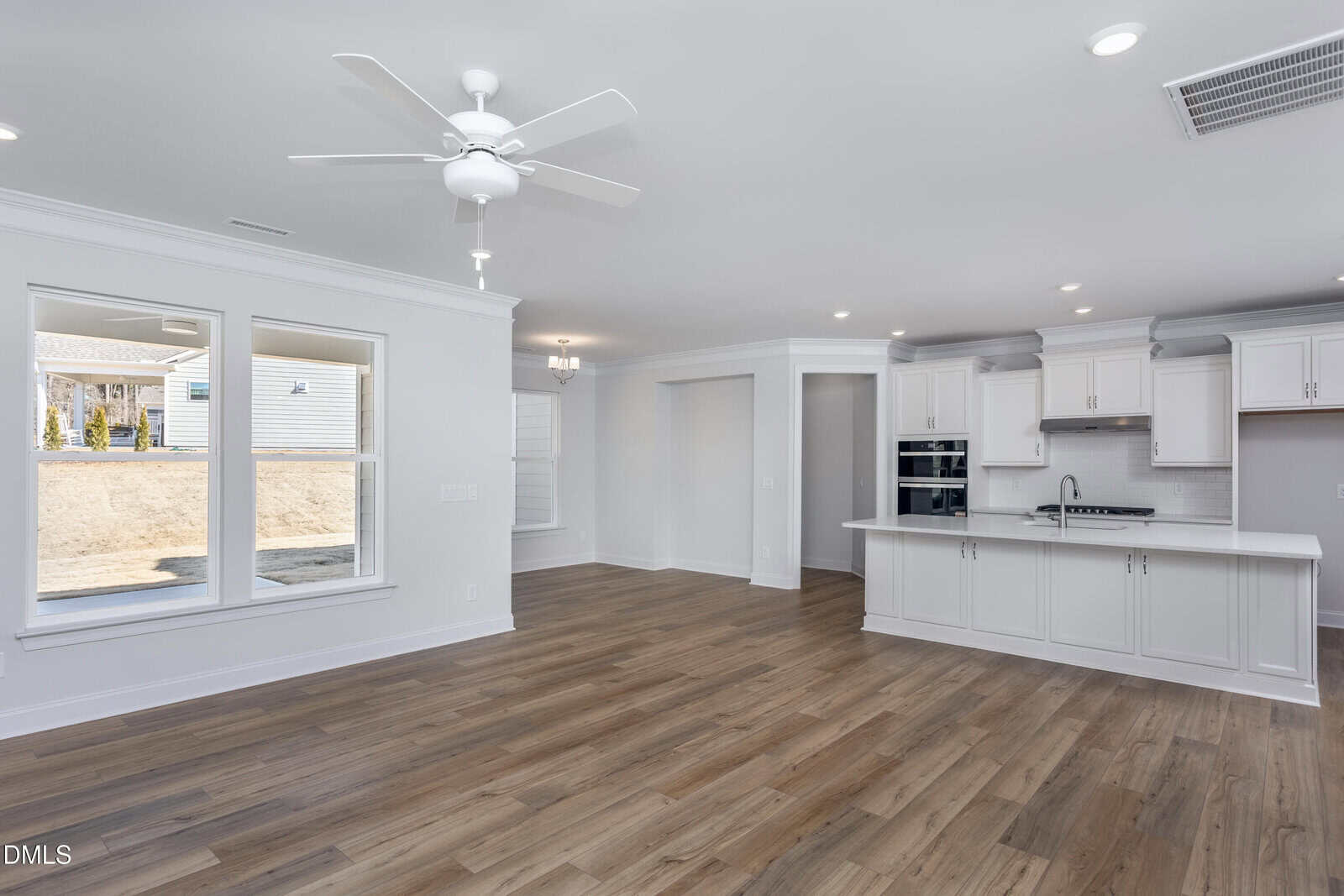 A bright, modern open-concept kitchen and living space with white cabinets, stainless steel appliances, light wood flooring, large windows, and a ceiling fan. Natural light fills the room.