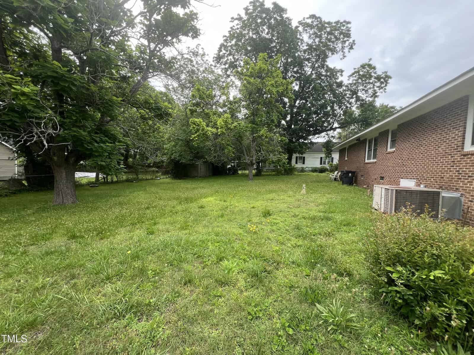 A grassy backyard with several large trees, bordered by a red brick house on the right. An air conditioning unit is next to the house, and a wooden fence is in the background. Sky is overcast.