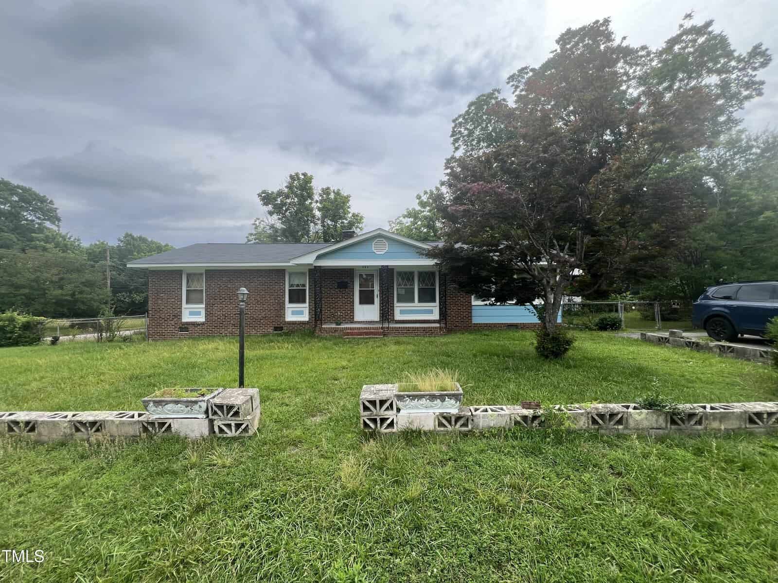 A single-story brick house with white trim and a front porch sits behind a lawn bordered by concrete blocks. A large tree stands to the right, and a blue vehicle is parked on the driveway. The sky is overcast.