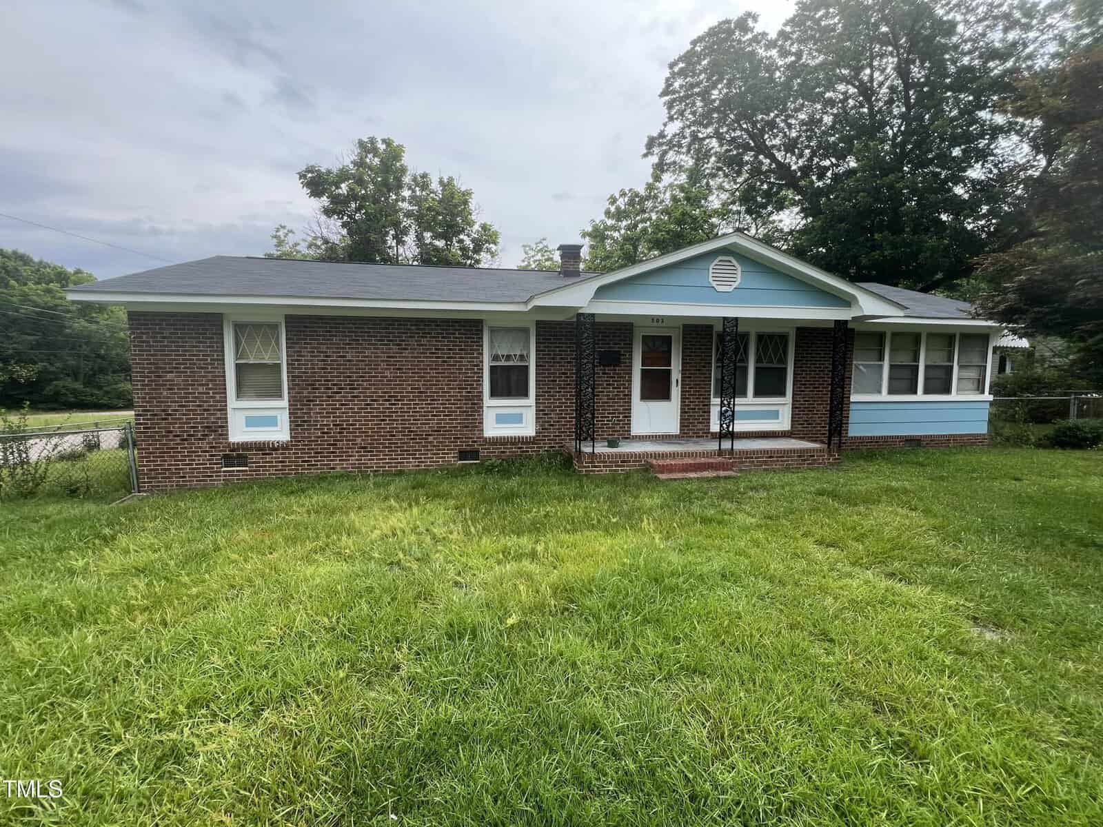 Charming single-story brick house at 503 Adams Street in Henderson, featuring a black roof, white trim, and blue accents on the porch and sunroom, set on a grassy lawn with trees in the background.