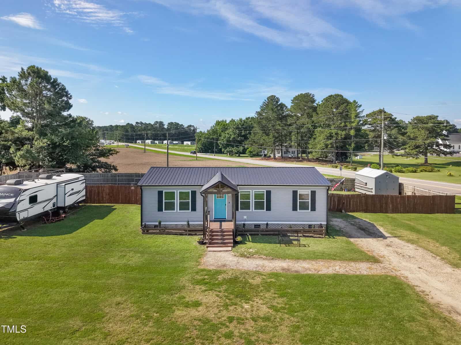 A small, single-story gray house with a blue front door, wooden steps, and a metal roof sits on a large grassy lot at 10 Wilders Lane in Louisburg. A driveway runs beside it; RVs and a wooden fence are nearby. Trees and a field are visible in the background.