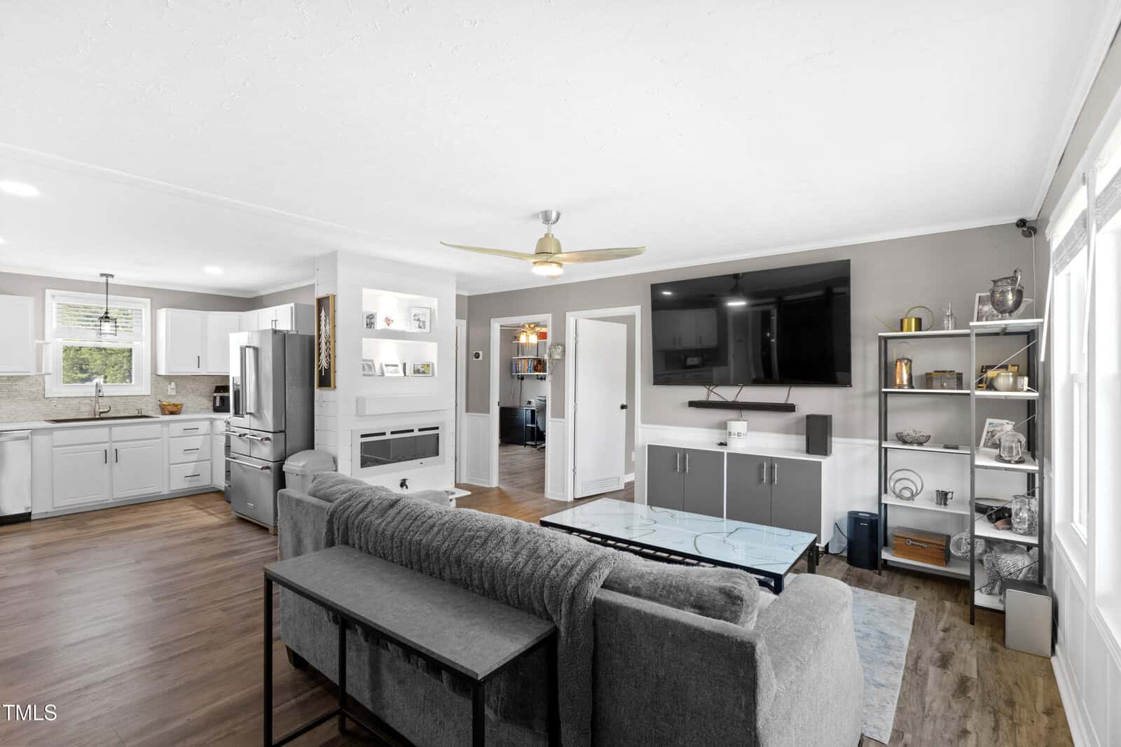 Open-concept living room and kitchen with gray sectional sofa, glass coffee table, wall-mounted TV, shelves, stainless steel appliances, and wood flooring. A small office is visible through an open doorway. Natural light fills the space.