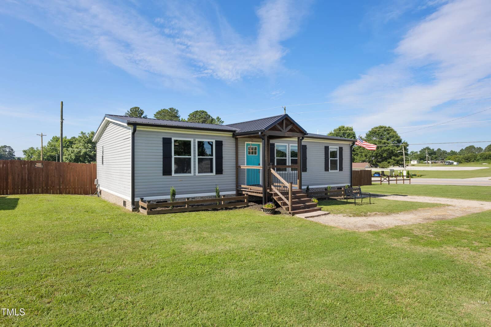 Single-story gray manufactured home with a teal front door and small covered porch, set on a grassy lot with a dirt driveway. An American flag is displayed on a wooden fence. Trees and a road are visible in the background.