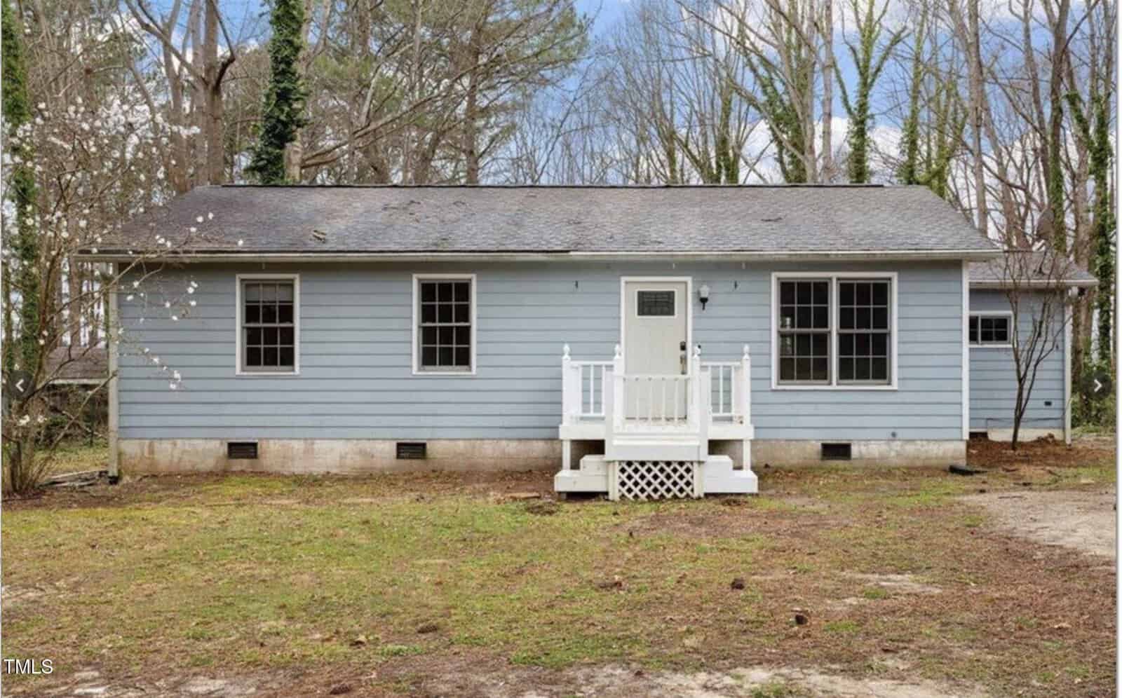A small, single-story blue house with white trim at 136 Dixie Lane, Youngsville, featuring a gray shingle roof and a cozy front porch with white railings, set in a yard with patchy grass and tall trees.