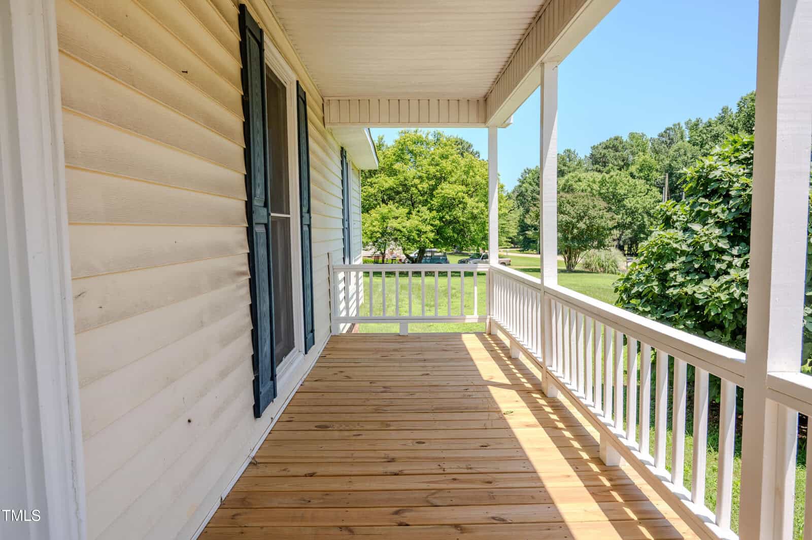 A wooden front porch with white railing and posts, attached to a light-colored house with a window on the left. Green trees and grass are visible in the background on a sunny day.