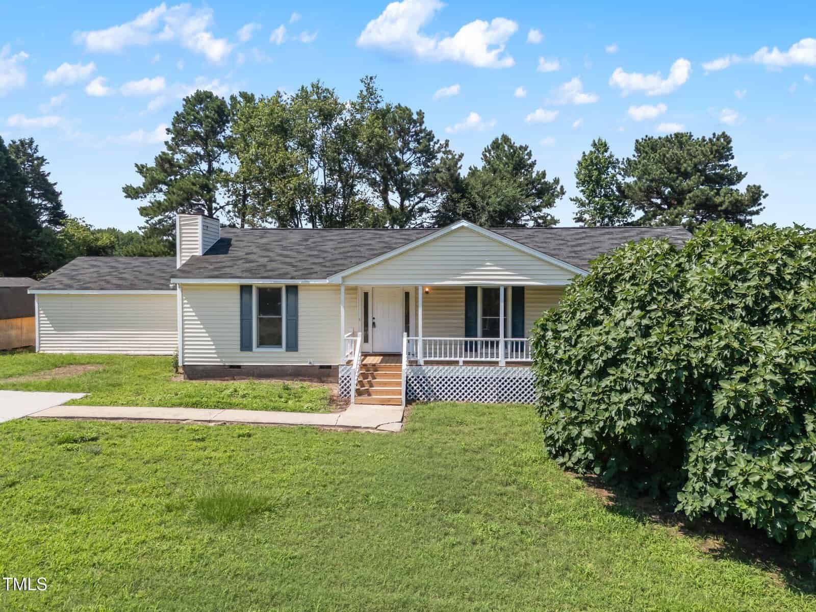Single-story house with light siding, dark roof, front porch, and steps leading to the entrance. Large green bush to the right and a grassy lawn with trees and a blue sky in the background. Concrete driveway on the left.