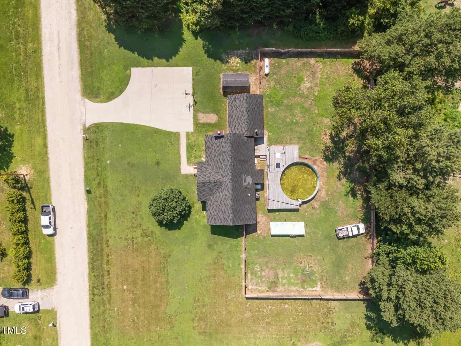 Aerial view of a house with a dark roof, fenced backyard, circular above-ground pool, driveway, two parked vehicles, and green lawn bordered by trees.