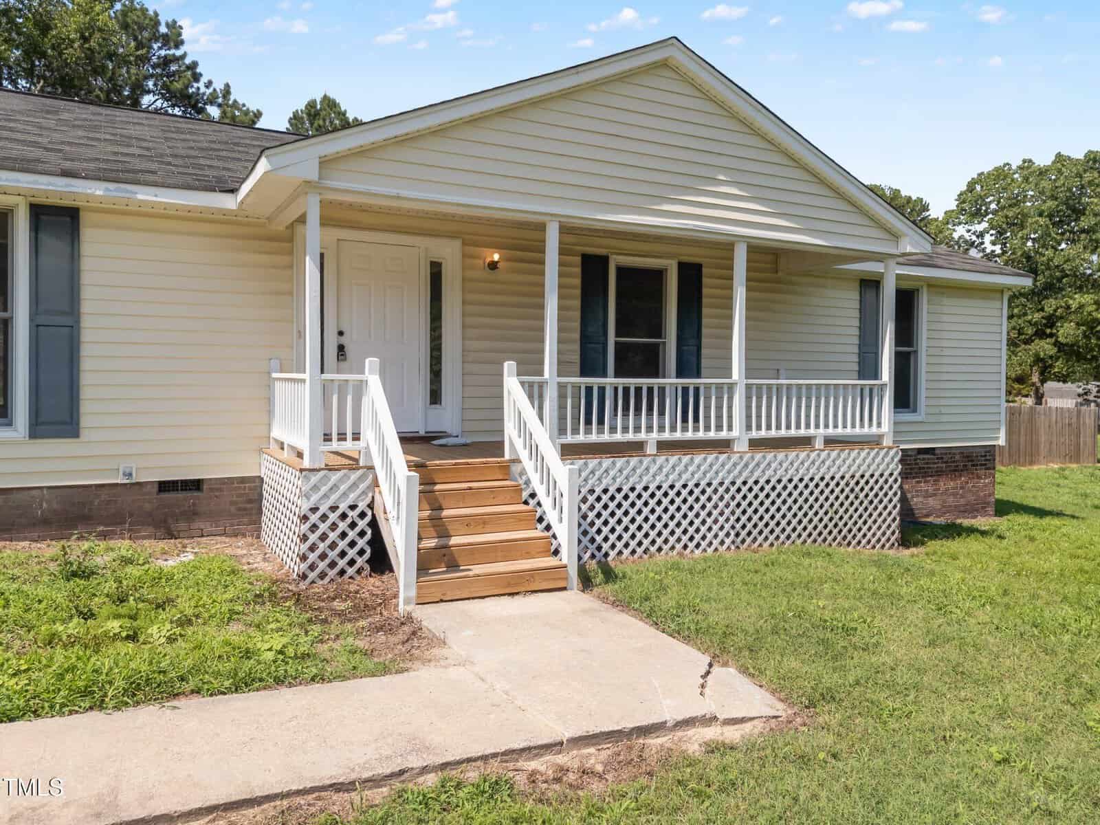 Single-story yellow house with white trim, a covered front porch, wooden steps, and a latticed porch base. There is a concrete walkway leading to the entrance, and a grassy yard surrounds the house. Trees are in the background.