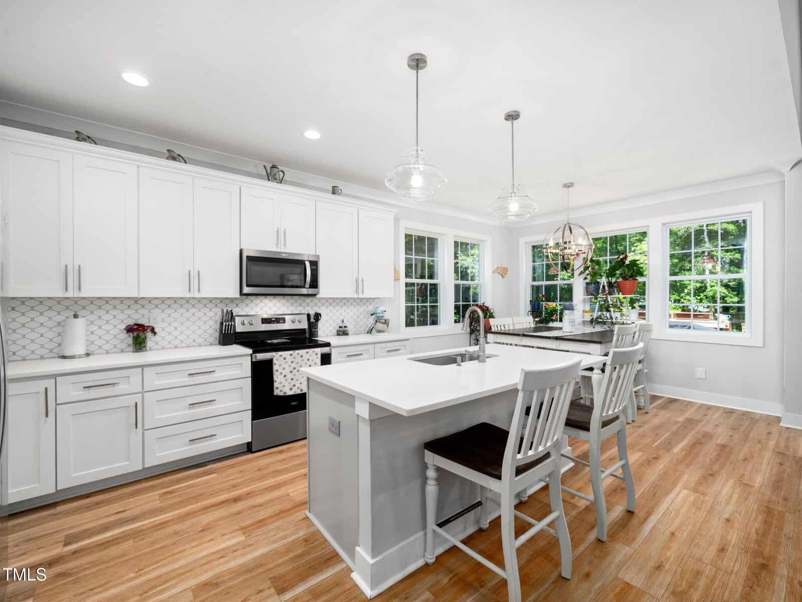 Modern kitchen with white cabinets, stainless steel appliances, a large island with two chairs, pendant lights, wooden floors, and large windows letting in natural light. A dining area is visible in the corner.