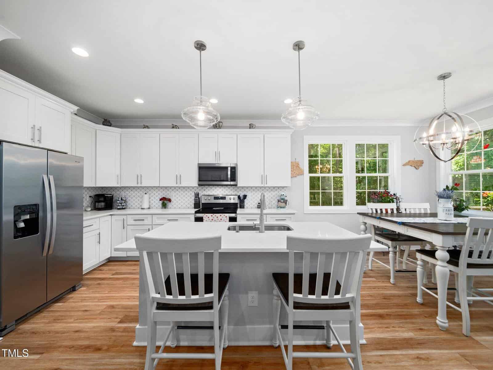Bright, modern kitchen with white cabinets, stainless steel appliances, an island with two white chairs, a dining table with chairs, pendant lighting, wood floors, and large windows letting in natural light.