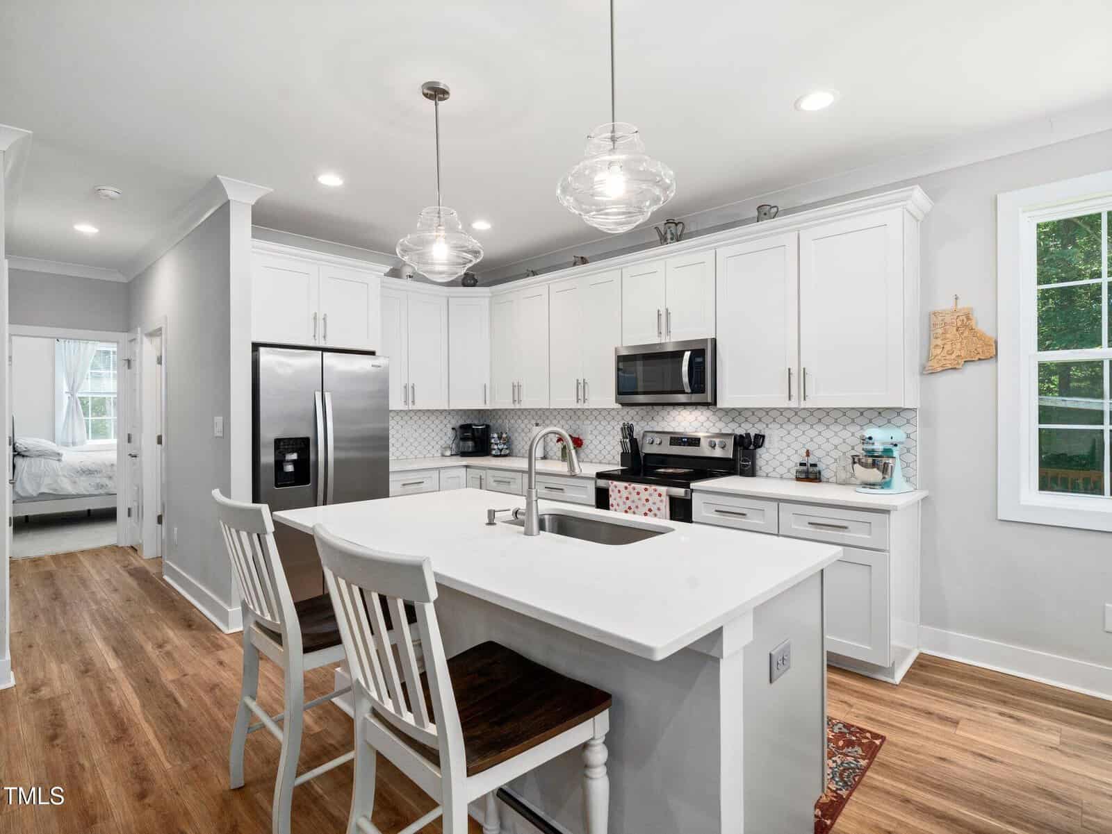 Bright, modern kitchen with white cabinets, stainless steel appliances, a central island with sink and two chairs, wood flooring, pendant lights, and a window letting in natural light. A bedroom is visible through an open doorway.