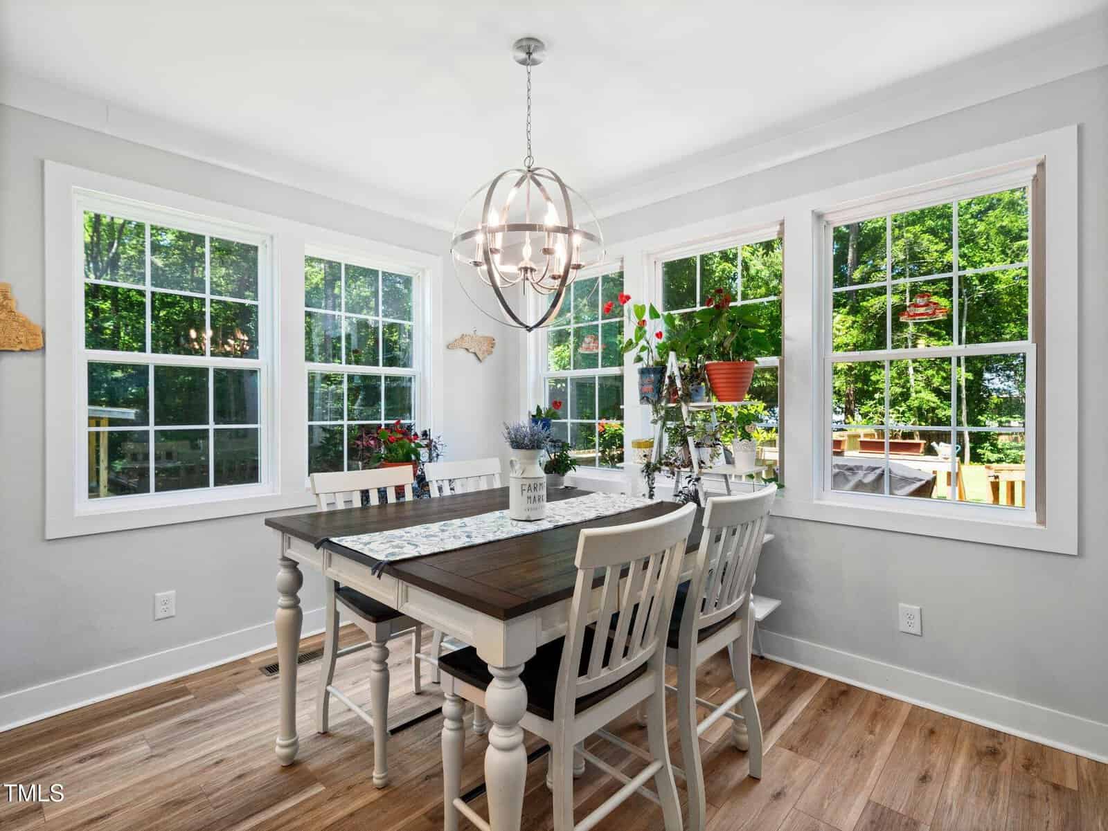 A bright dining area with a wooden table and six white chairs, set near large windows overlooking a green yard. A modern chandelier hangs above, and houseplants decorate the windowsill. Light wood floors complete the look.
