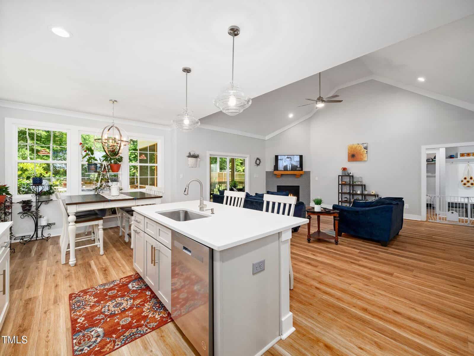 Open-concept kitchen and living area with wood floors, white cabinets, a center island, dining table by large windows, blue sofas, ceiling fans, and a TV on the far wall. Natural light fills the space.