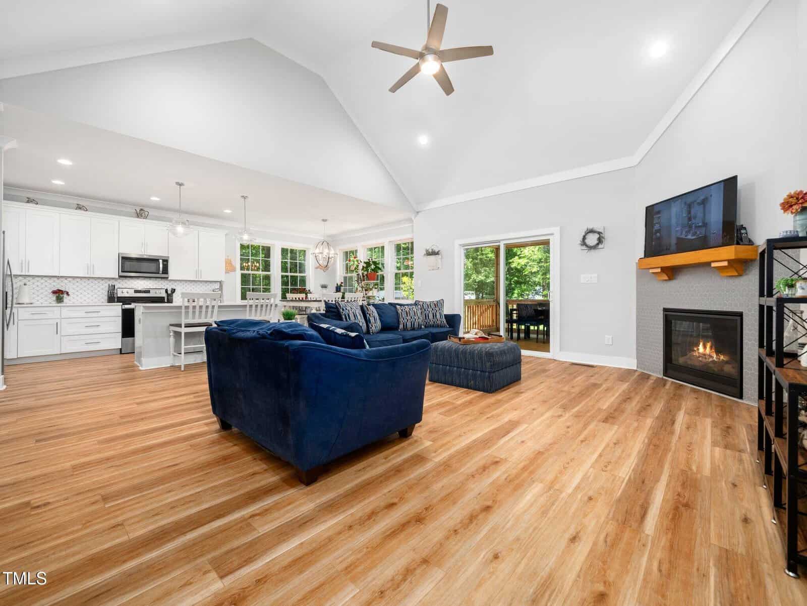 Open living space with wood floors features a navy sectional, fireplace with mounted TV, kitchen with white cabinets and island, dining area, vaulted ceiling, and ceiling fan. Sliding doors lead to an outdoor area.
