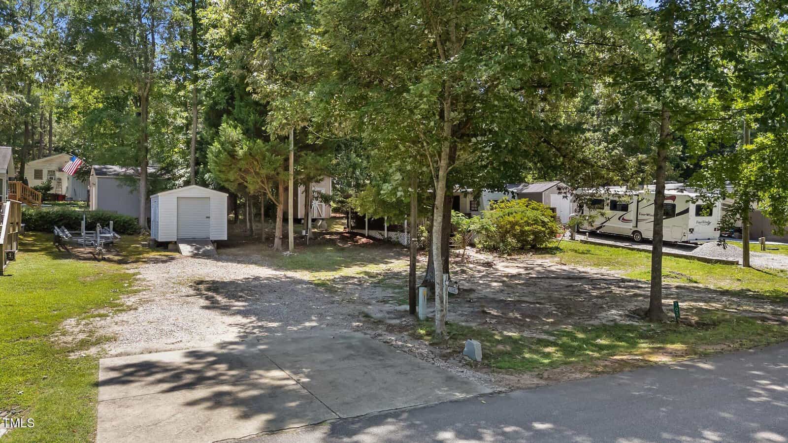 A vacant lot with a concrete driveway, gravel area, and a small shed. Mature trees provide shade, and neighboring RVs and homes are visible. The area is clean and surrounded by greenery.