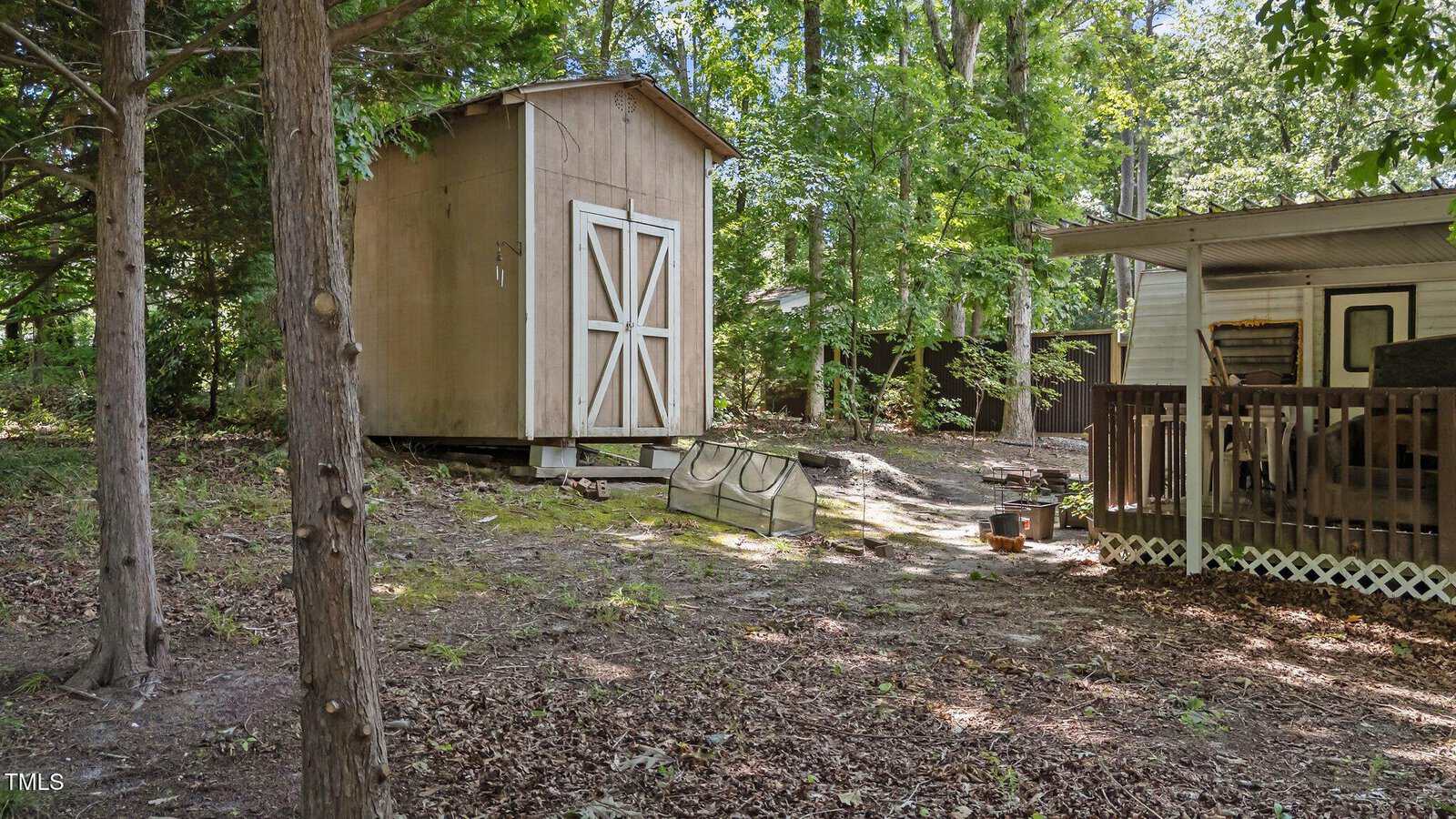 A wooden shed with double doors sits on a slightly raised foundation near trees. To the right, a covered porch with a railing is attached to a house. The ground is covered with dry leaves and patches of dirt.