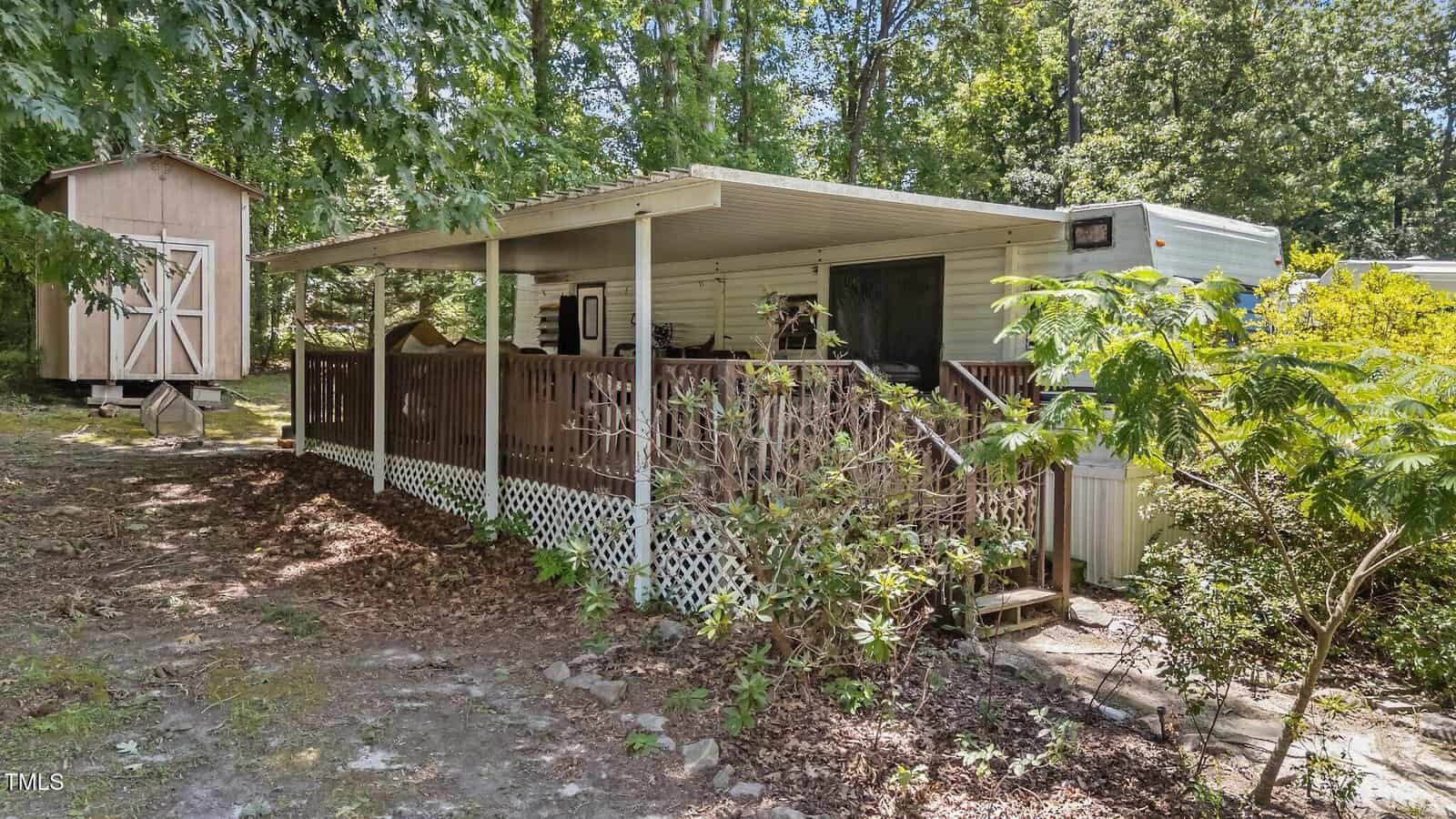 A small white mobile home with a covered wooden porch and railing, surrounded by trees and bushes, is nestled on 103 Big Horn Drive in Louisburg. A wooden shed sits in the background on the left, while dirt and leaves cover the ground around the porch area.