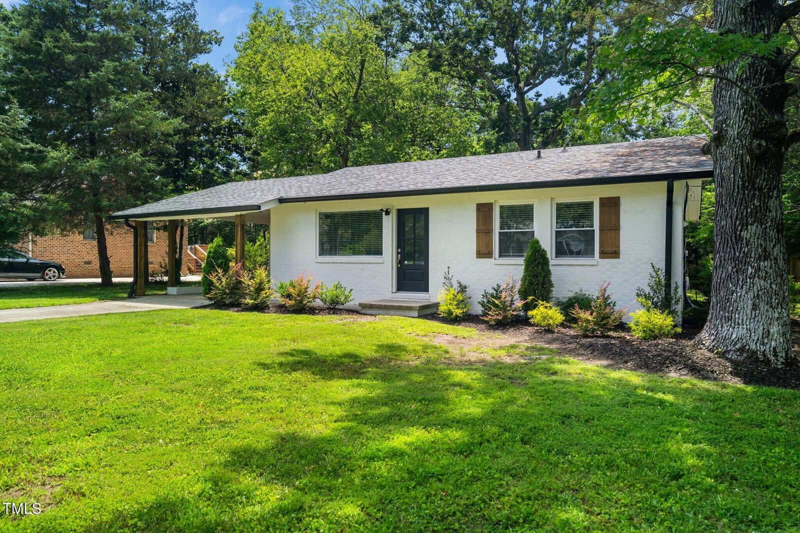 A small, single-story white brick house at 313 13th Street, Butner, with a dark front door, wooden shutters, a covered carport, and neatly trimmed shrubs in front, surrounded by green grass and tall trees.