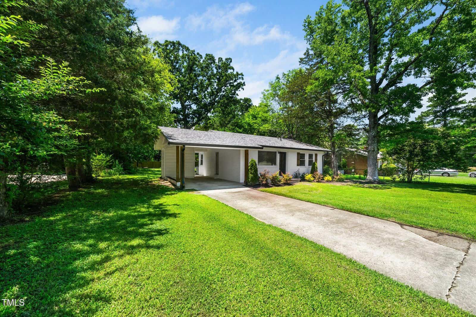 A single-story house with light-colored siding, a carport, and a concrete driveway, surrounded by green grass, trees, and landscaping under a partly cloudy sky.