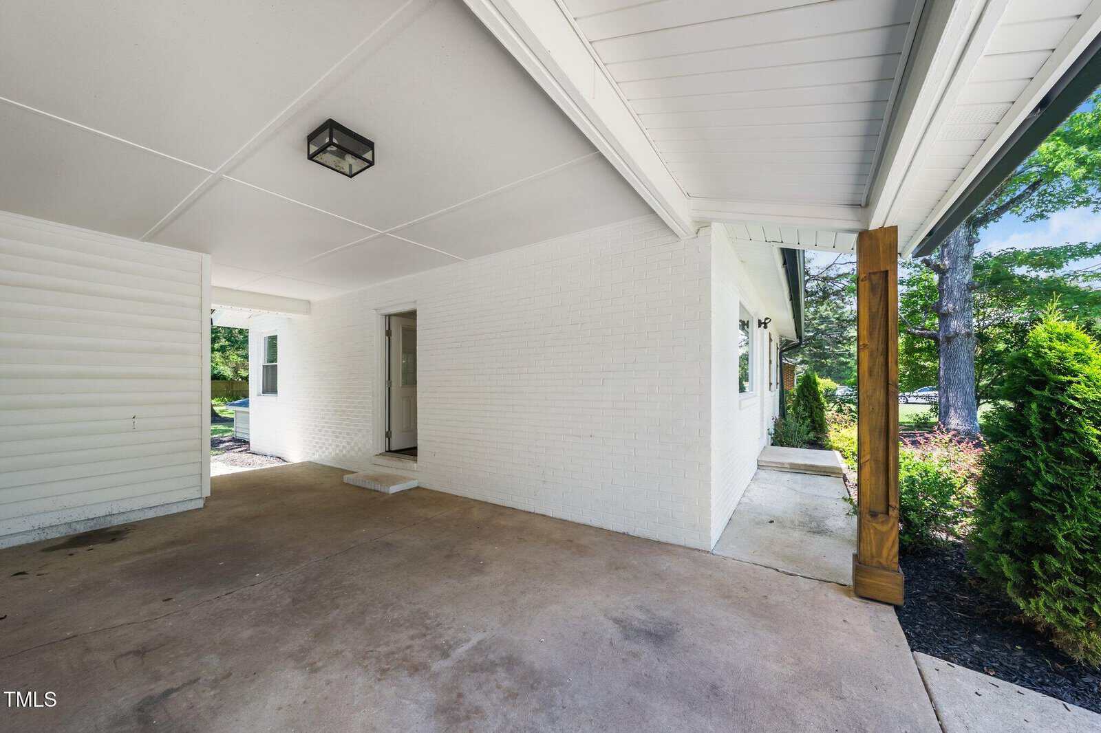 Covered carport area attached to a white brick house with a single door entry, concrete flooring, wooden support post, and some green shrubs visible along the perimeter.