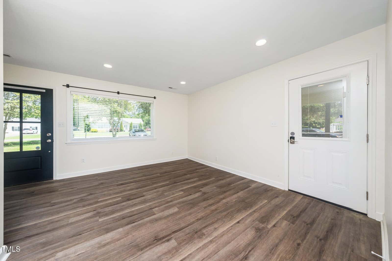 Bright, empty room with light walls, recessed lighting, wood-style flooring, a large window with blinds, and two white doors, one with glass, providing views of a suburban street and greenery outside.