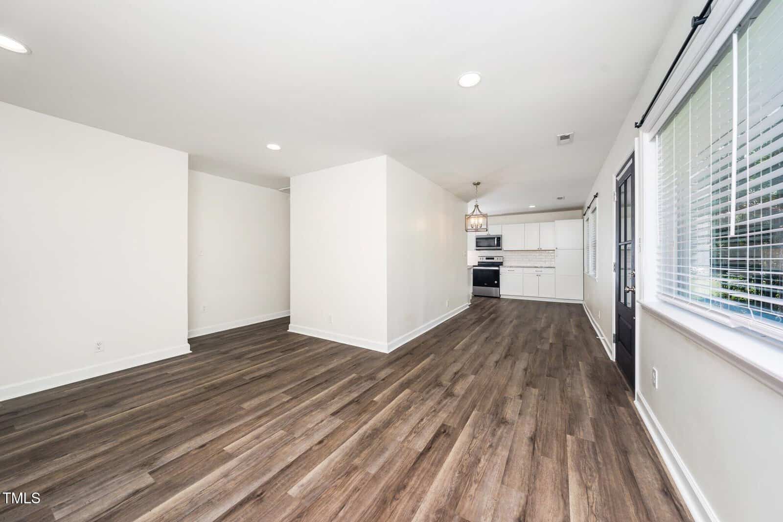 A spacious open-concept living area with wood-look flooring, white walls, recessed lighting, and large windows. The kitchen in the background features white cabinets, a stove, and a center light fixture.