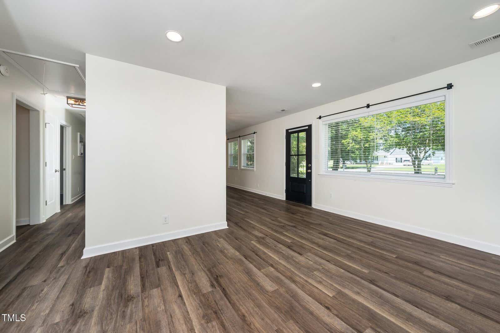 A spacious, empty room with white walls, wood-style flooring, large windows with blinds, a black door, and recessed ceiling lights. Natural light enters through the windows, and an open hallway leads to other rooms.