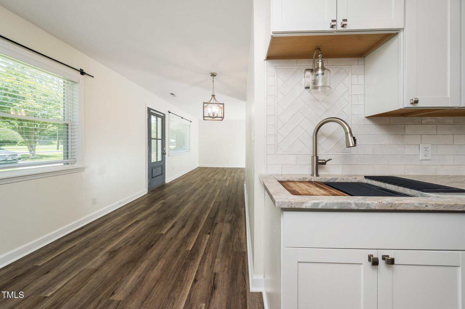 View from a kitchen with white cabinets and a modern sink, looking into an open living area with wood flooring, large window, dark-colored door, and a geometric light fixture hanging from the ceiling.