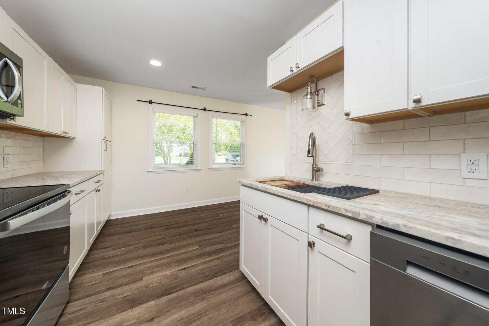 Modern kitchen with white cabinets, stainless steel appliances, light marble countertops, subway tile backsplash, and two large windows letting in natural light. Dark wood floors contrast with the bright, clean space.