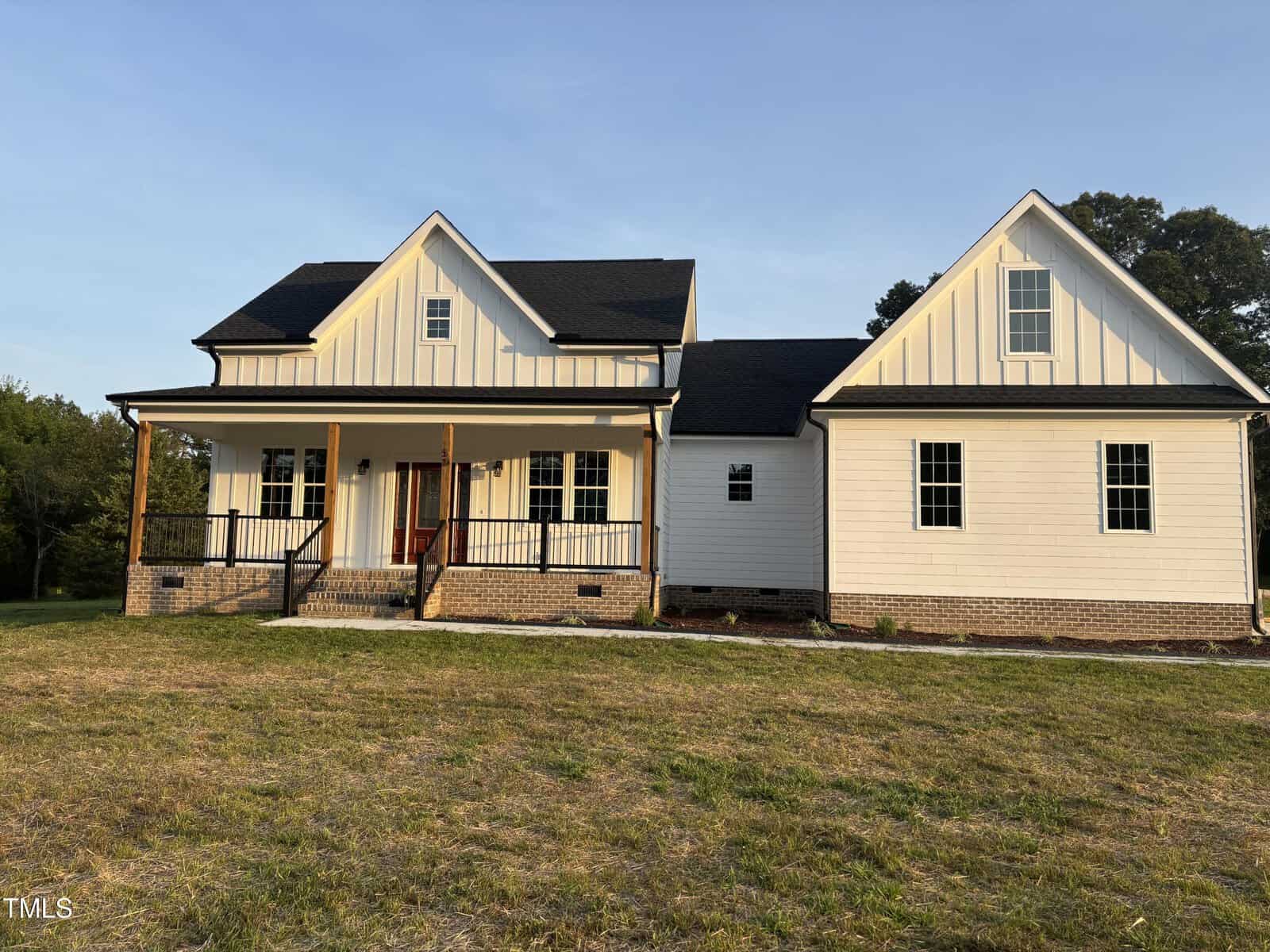 A two-story modern farmhouse with white board and batten siding, a black roof, covered front porch with wooden columns, and brick steps, set on a grassy yard during daylight.