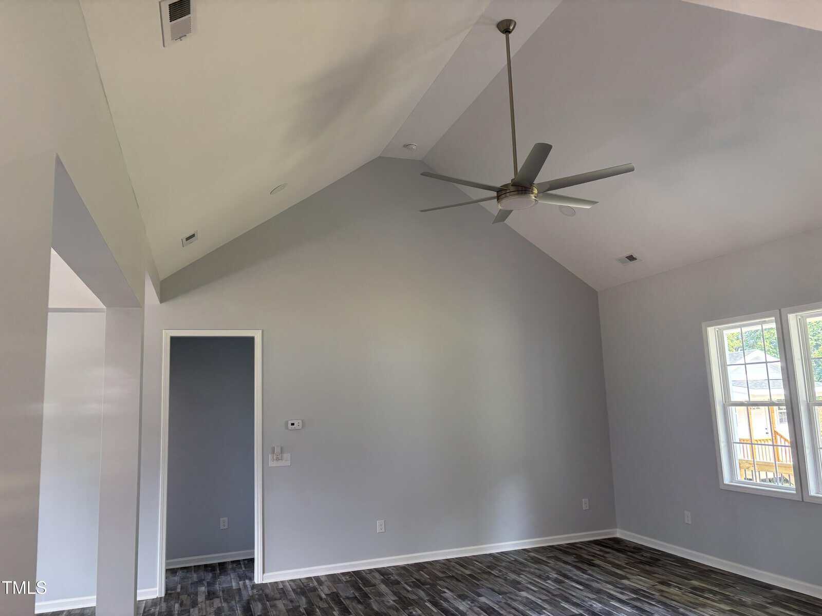 A room with light gray walls, vaulted ceiling, ceiling fan, large window, and dark wood-look flooring. There is a small doorless opening and a thermostat on one wall. Natural light enters from the window.
