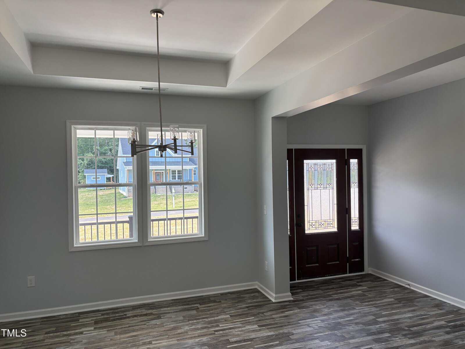 A bright room with gray walls, tray ceiling, modern light fixture, wood-style flooring, large window, and a dark front door with glass panels. Outside, a blue house and grassy yard are visible through the window.
