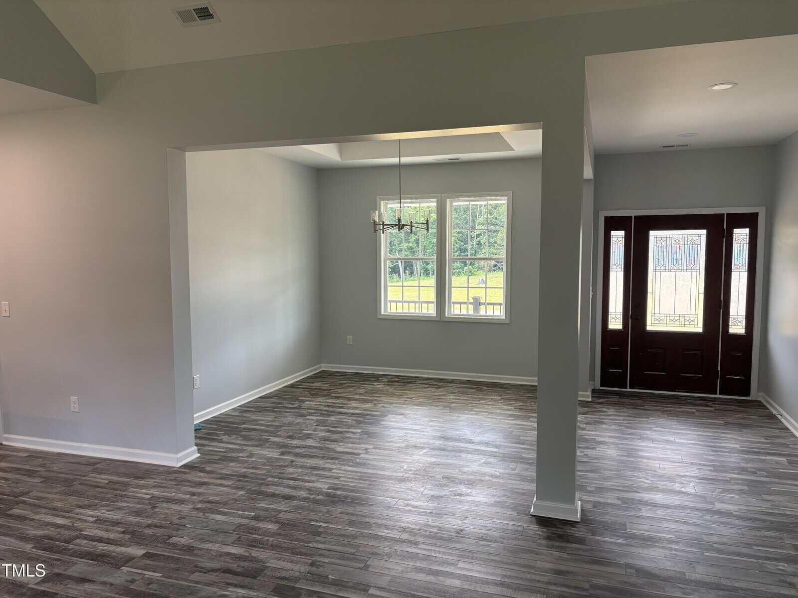 A spacious, empty room with gray walls, dark wood flooring, large windows letting in natural light, a modern chandelier, and a double front door with glass panels.