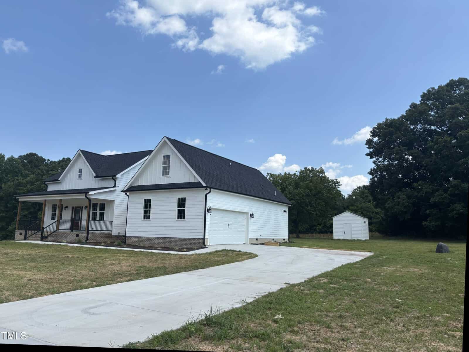 A white, single-story house with black roof and attached garage sits on a grassy lot. A long concrete driveway leads to the house, and a small white shed is visible in the background under a partly cloudy sky.
