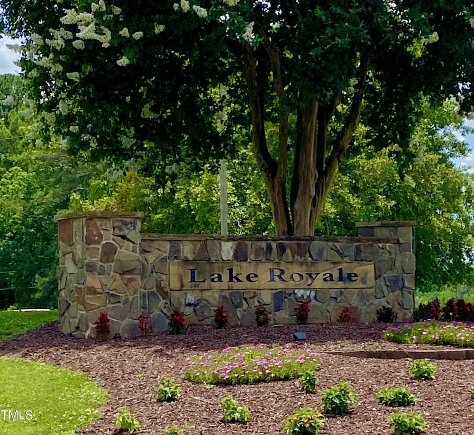 A stone sign with the text "Lake Royale" stands in front of a landscaped area with mulch, small plants, and a large tree in the background. The scene is outdoors with greenery and blue sky visible.