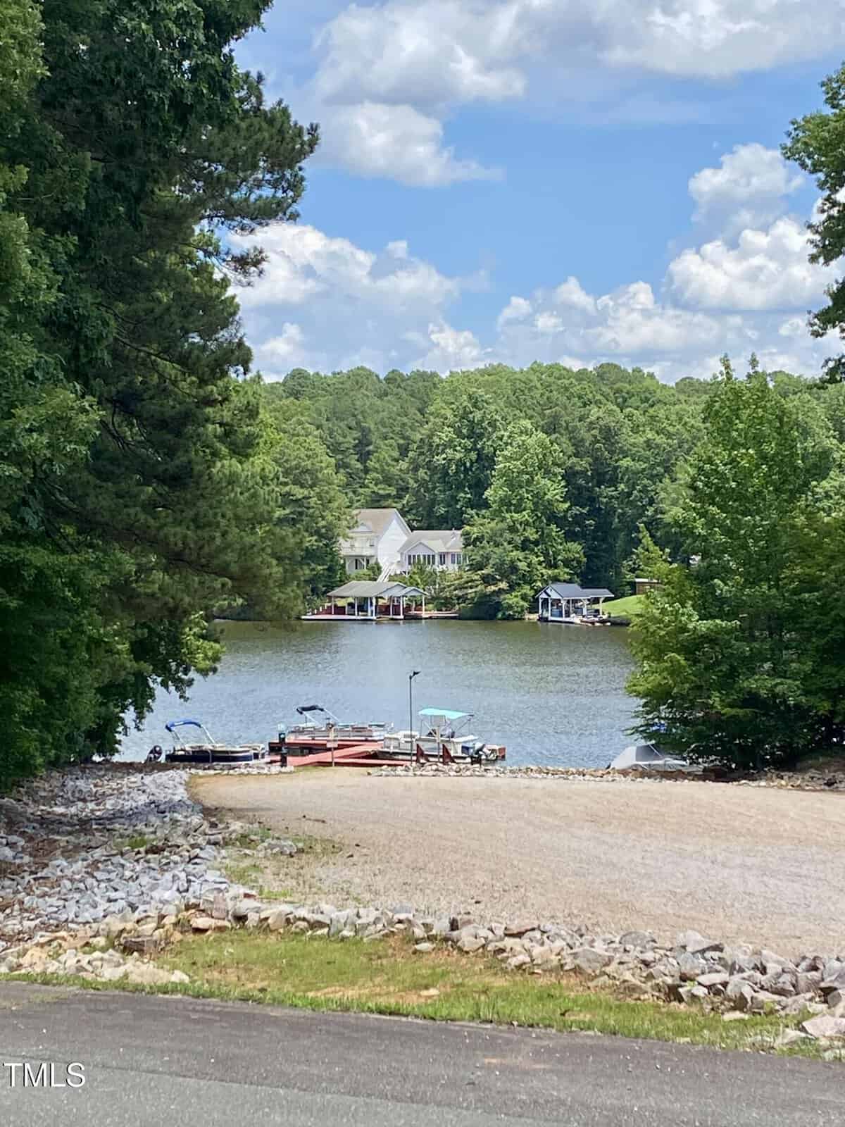 A gravel boat ramp at 106 Cherokee Drive in Louisburg leads down to a small lake with boats docked at the edge. Trees surround the water, and a house is visible among them across the lake under a partly cloudy sky.