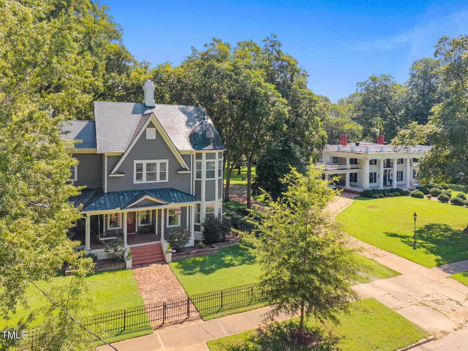 A two-story house with a covered front porch, gray siding, and white trim stands next to a wide driveway with a large white columned building in the background, surrounded by green lawns and mature trees.