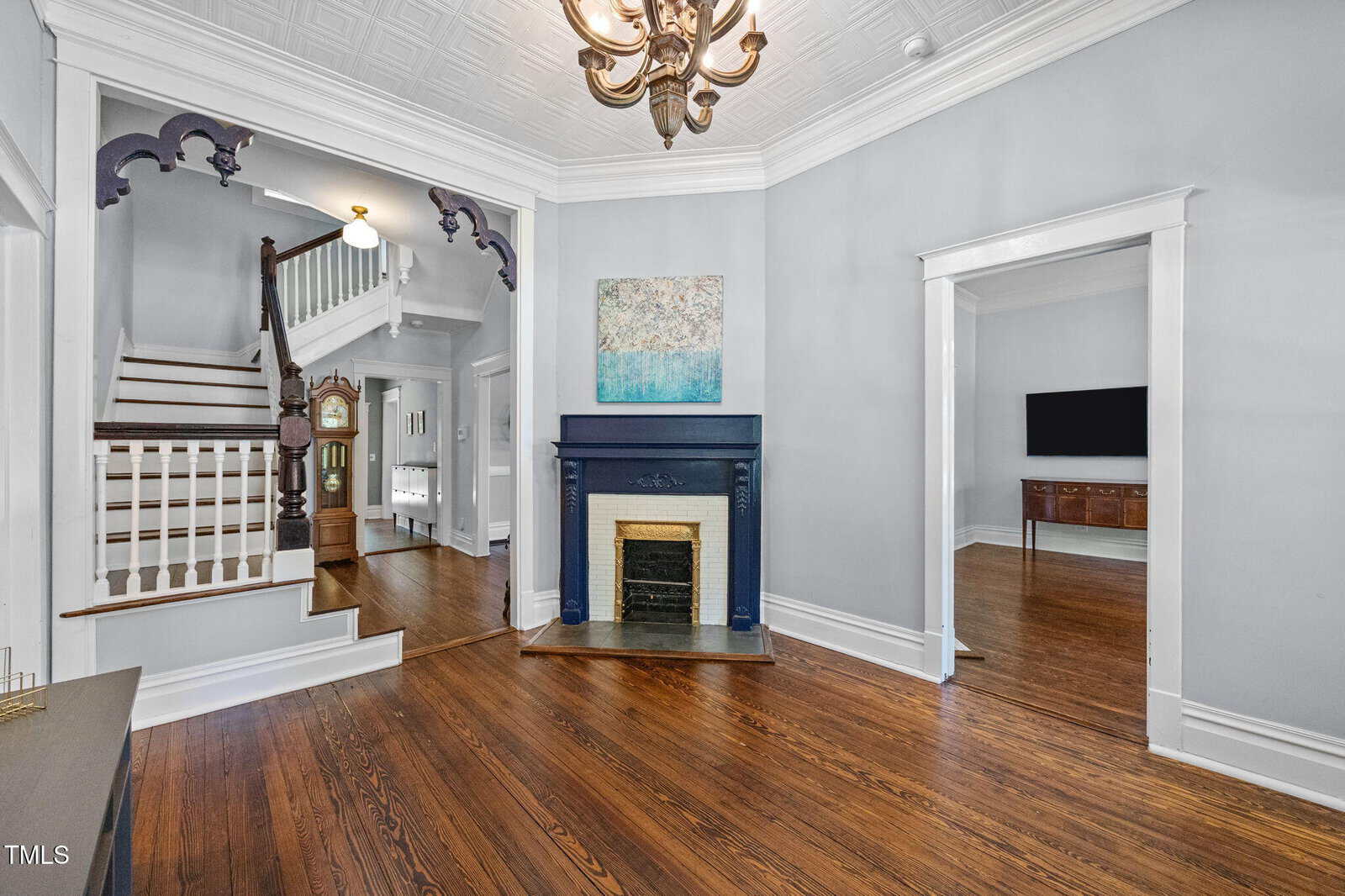 A living room with hardwood floors, a white ceiling with a decorative pattern, a chandelier, a dark blue fireplace with artwork above it, and a staircase with white spindles leading upstairs.