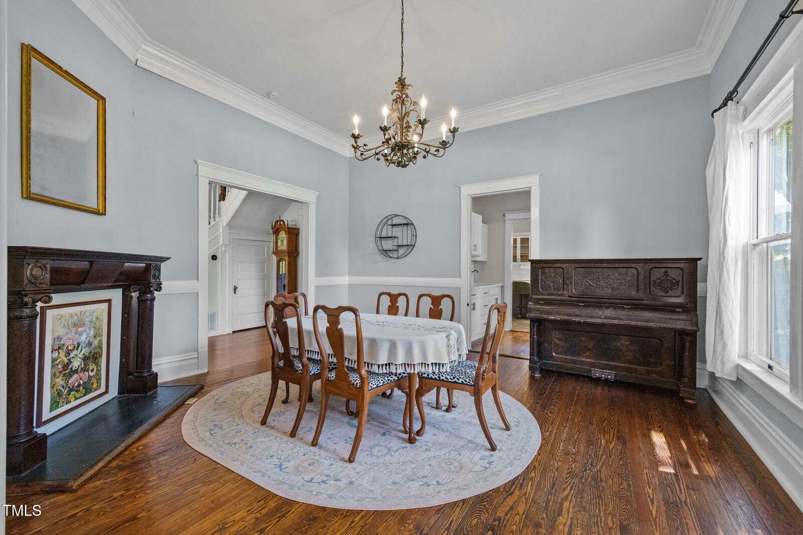 A dining room with a round table covered by a lace tablecloth, six wooden chairs, a chandelier, hardwood floors, a fireplace, a piano, and large windows letting in natural light.