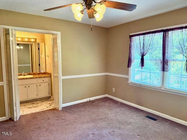 A bedroom with tan walls, white trim, and brown carpet. There is a ceiling fan with lights, a window with sheer purple curtains, and an open door leading to a bathroom with a vanity and mirror.
