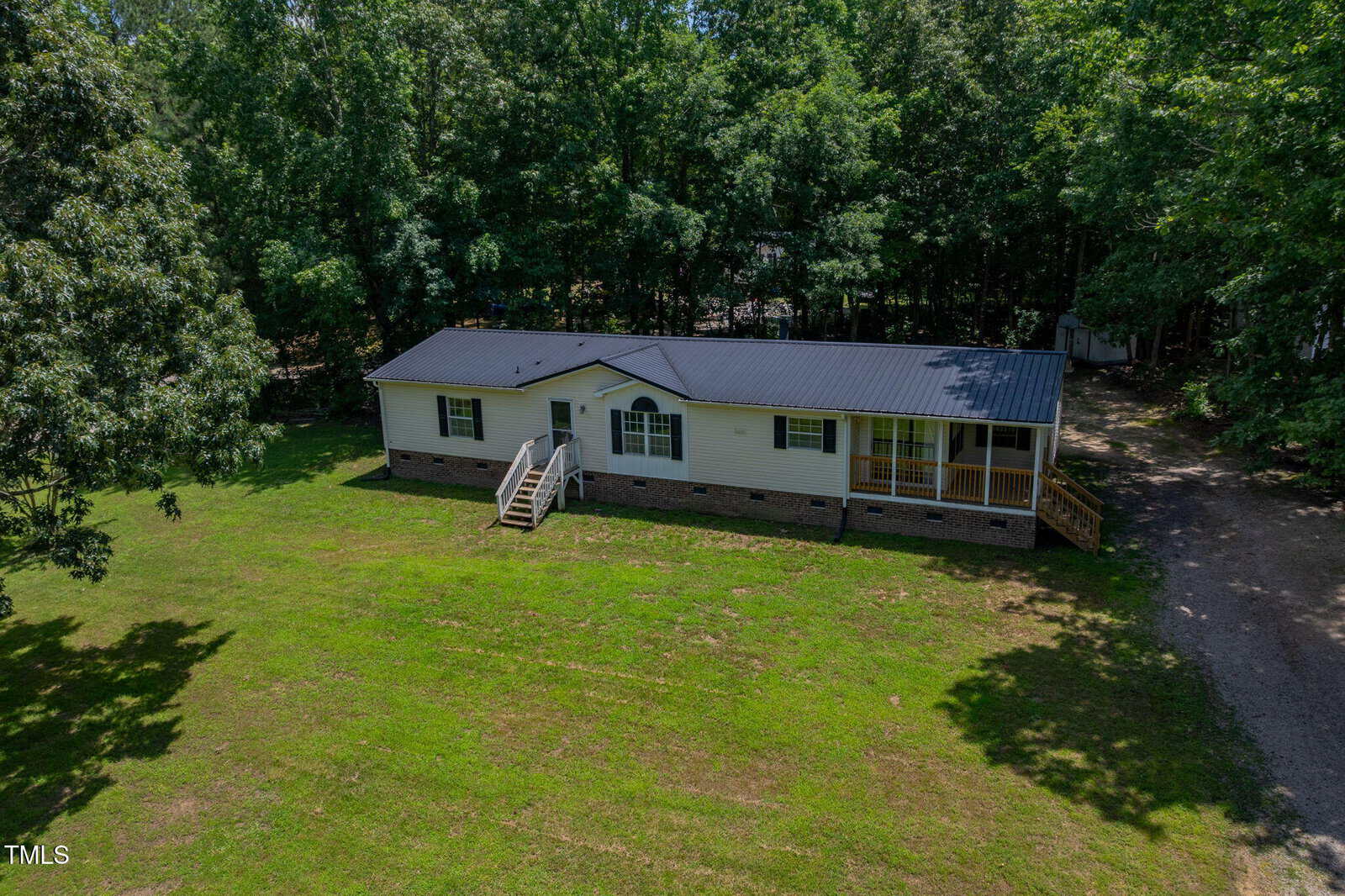 A single-story manufactured home with a dark roof, cream-colored siding, and brick foundation sits on a grassy lot surrounded by trees. There are front steps, a small porch, and a driveway on the right side.