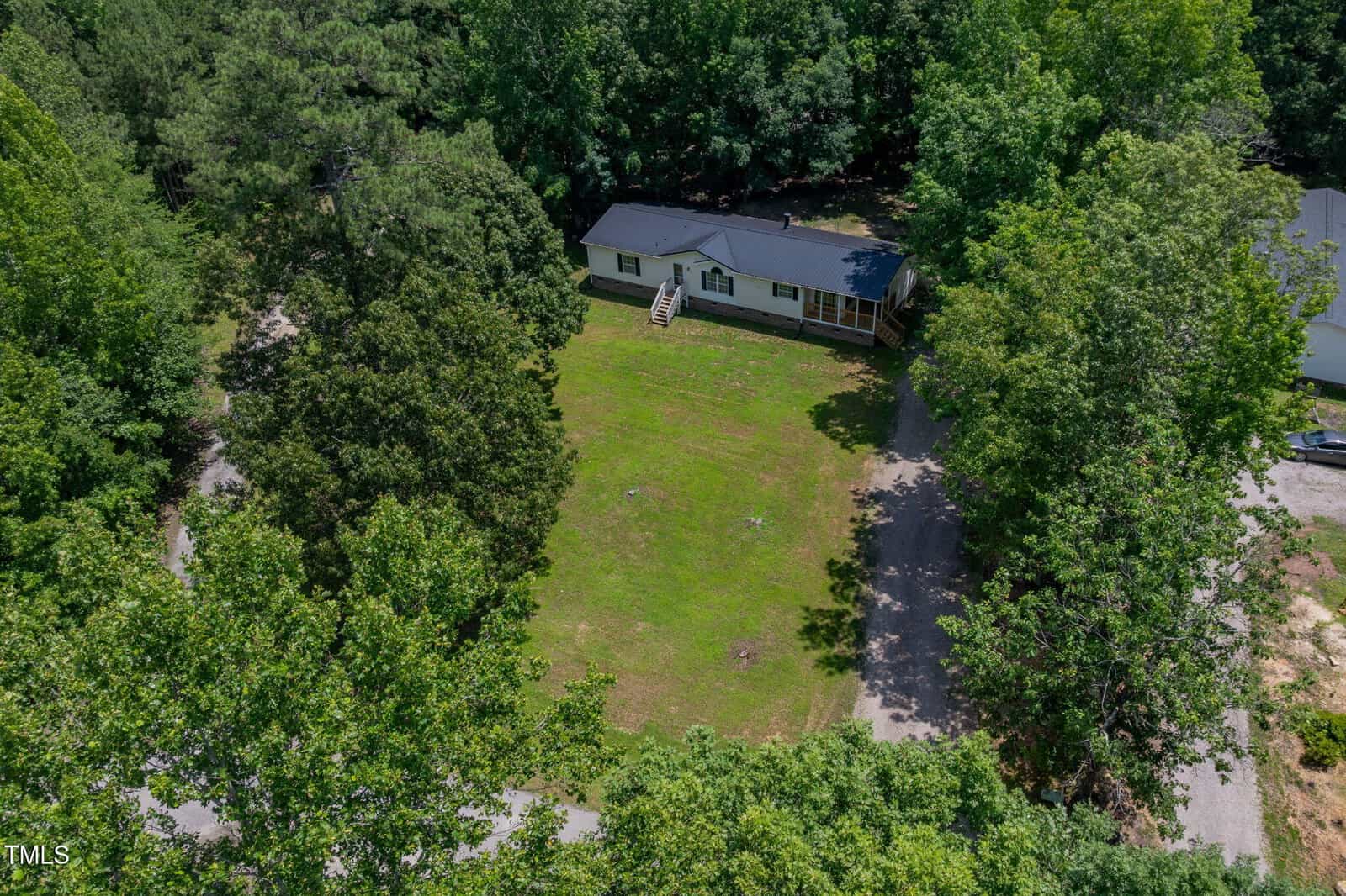 Aerial view of a single-story house with a dark roof and front steps, situated on a large grassy lawn surrounded by dense green trees and a gravel driveway.