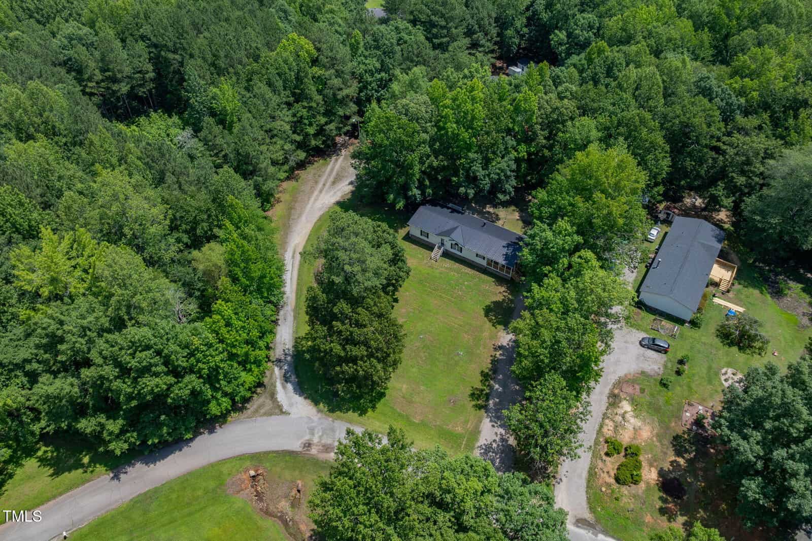 Aerial view of two homes surrounded by dense trees, each with a driveway leading to the street. The houses are separated by a large grassy area and patches of woodland. Several vehicles are parked outside one of the homes.