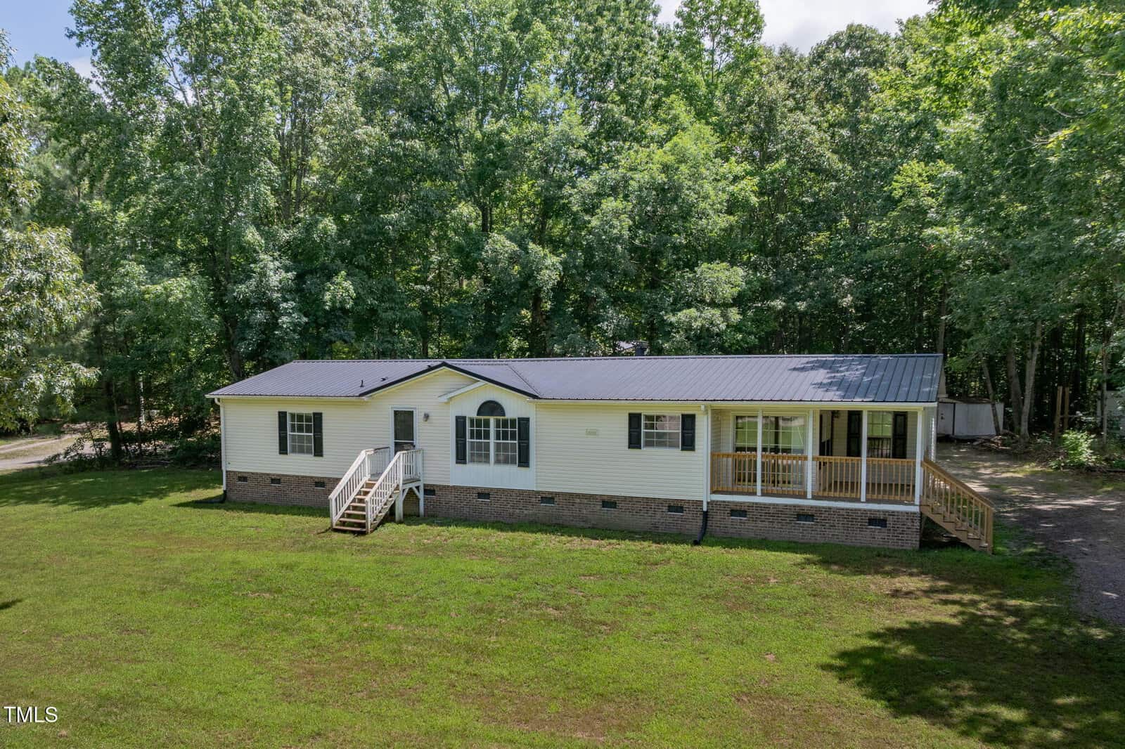 A single-story manufactured home at 40 Raccoon Crossing in Henderson features a dark metal roof, white siding, brick foundation, front steps, and a covered porch on a grassy lot surrounded by tall trees.