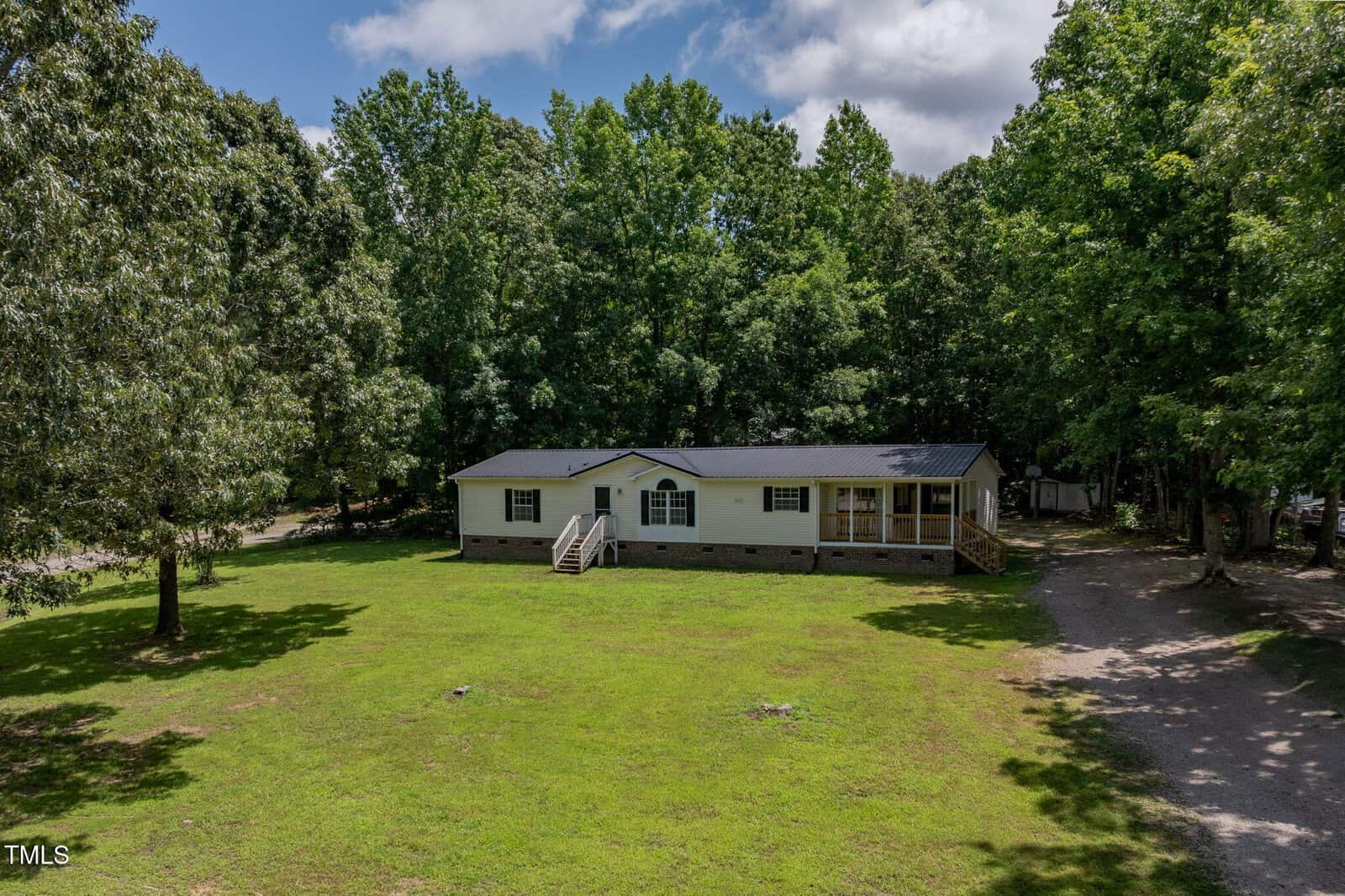 A single-story manufactured home with a covered front porch sits on a large grassy lot surrounded by trees. A gravel driveway leads up to the house, and sunlight filters through partly cloudy skies.