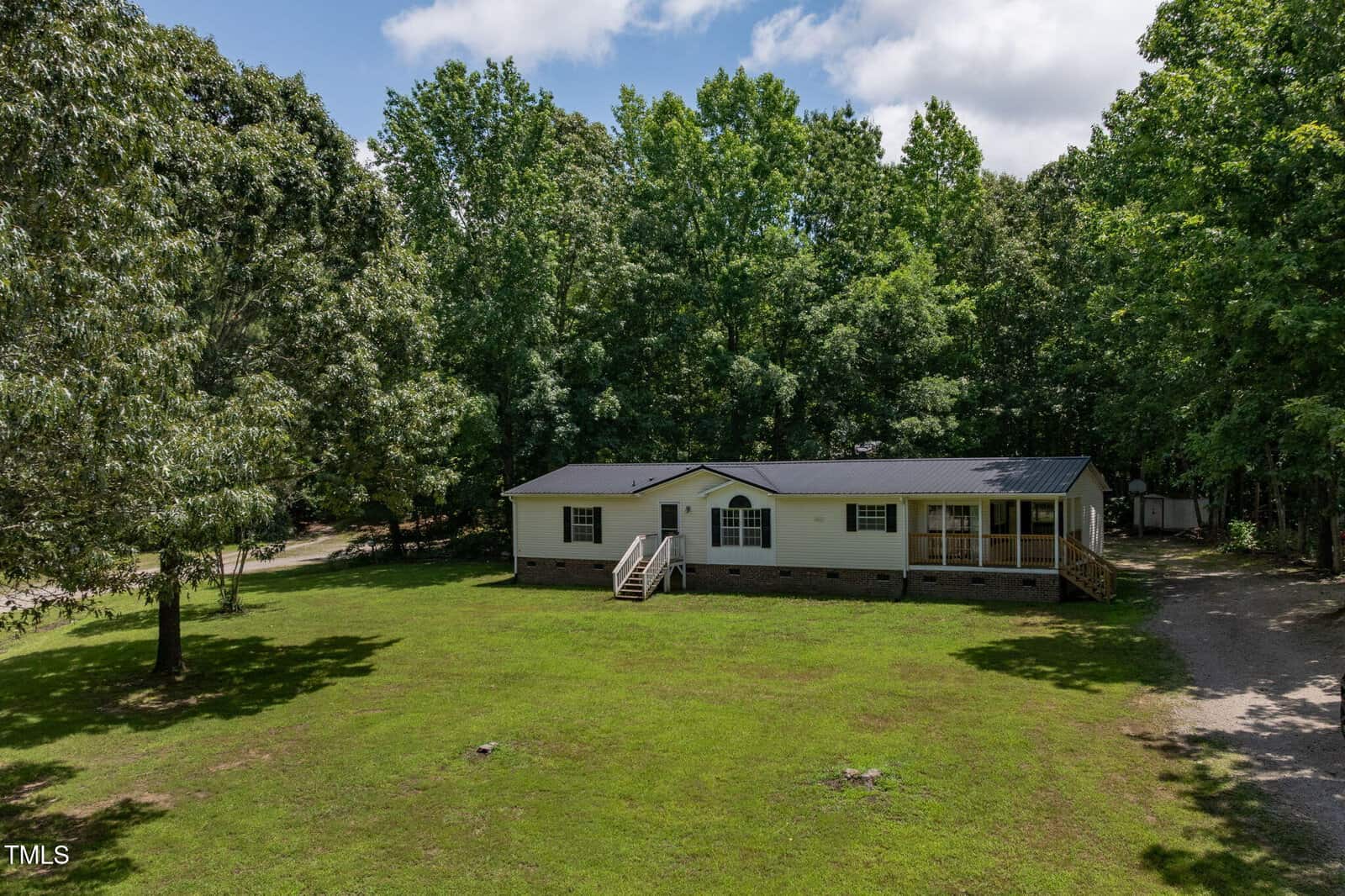 A single-story manufactured home with light-colored siding and a dark roof sits on a large grassy lawn, surrounded by tall, leafy trees under a partly cloudy sky. A wooden porch leads to the front entrance.